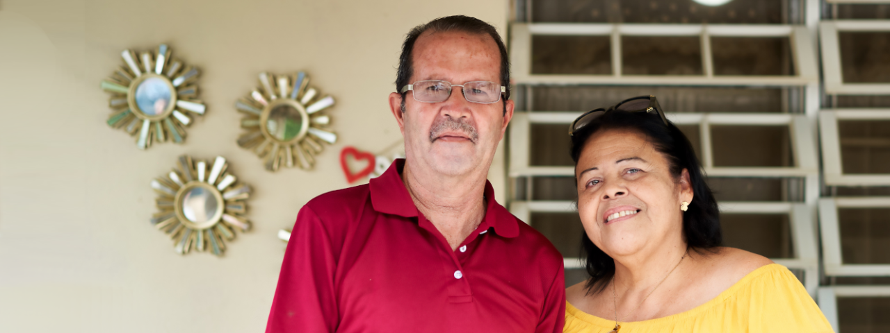 Pareja con camisas roja y amarilla, sonriendo frente a su hogar.